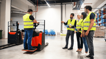 Workers in a warehouse wearing high-visibility vests observe a colleague operating an electric pallet jack during a hands-on safety training demonstration.